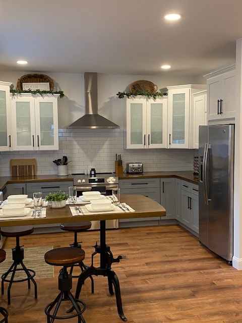 The kitchen features modern cabinetry with glass door accents and a spacious layout. A stainless steel refrigerator stands prominently, complemented by a gas stove and stylish exhaust hood. A wooden dining table is set for four, showcasing dinnerware. Warm wood flooring completes the inviting atmosphere.