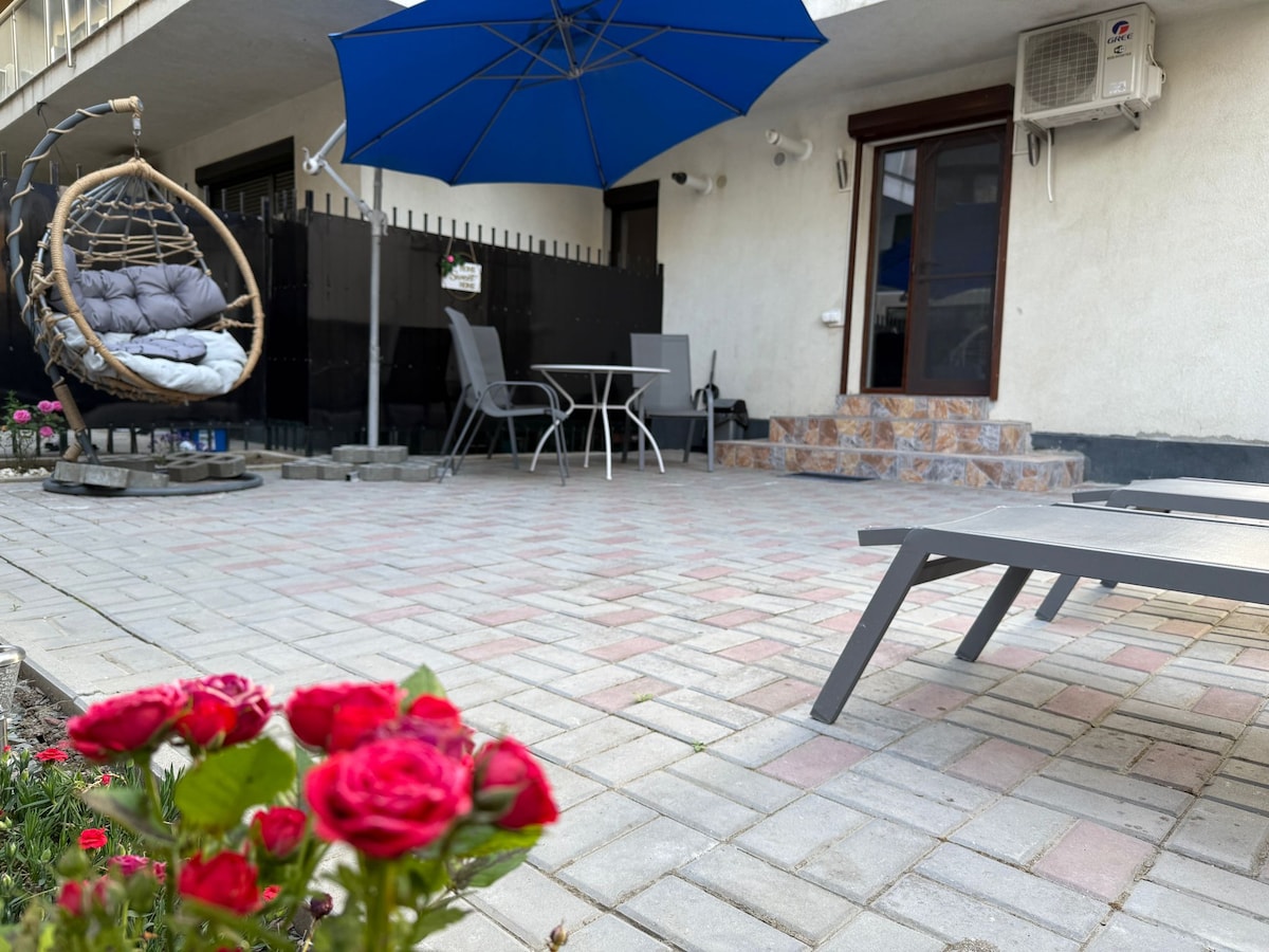 A private terrace is highlighted, featuring a hanging chair and a table with chairs under a blue umbrella. Sun loungers are positioned nearby, while vibrant red roses add color to the edges of the stone-paved area.