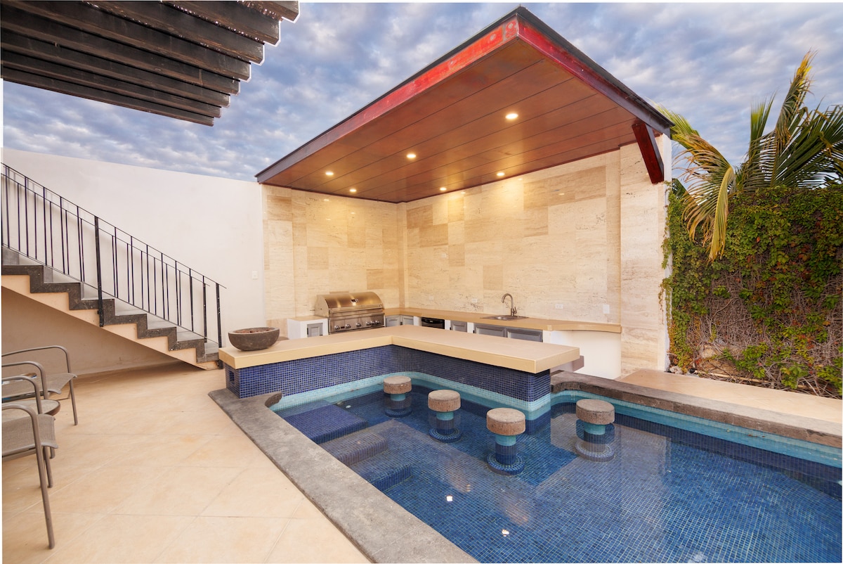 An outdoor kitchen area is visible beside a heated pool, featuring a stone countertop, grill, and sink. Steps lead to an upper level. The pool is framed by blue tiles, and seating is provided by rounded stone stools. Lush greenery borders the space.