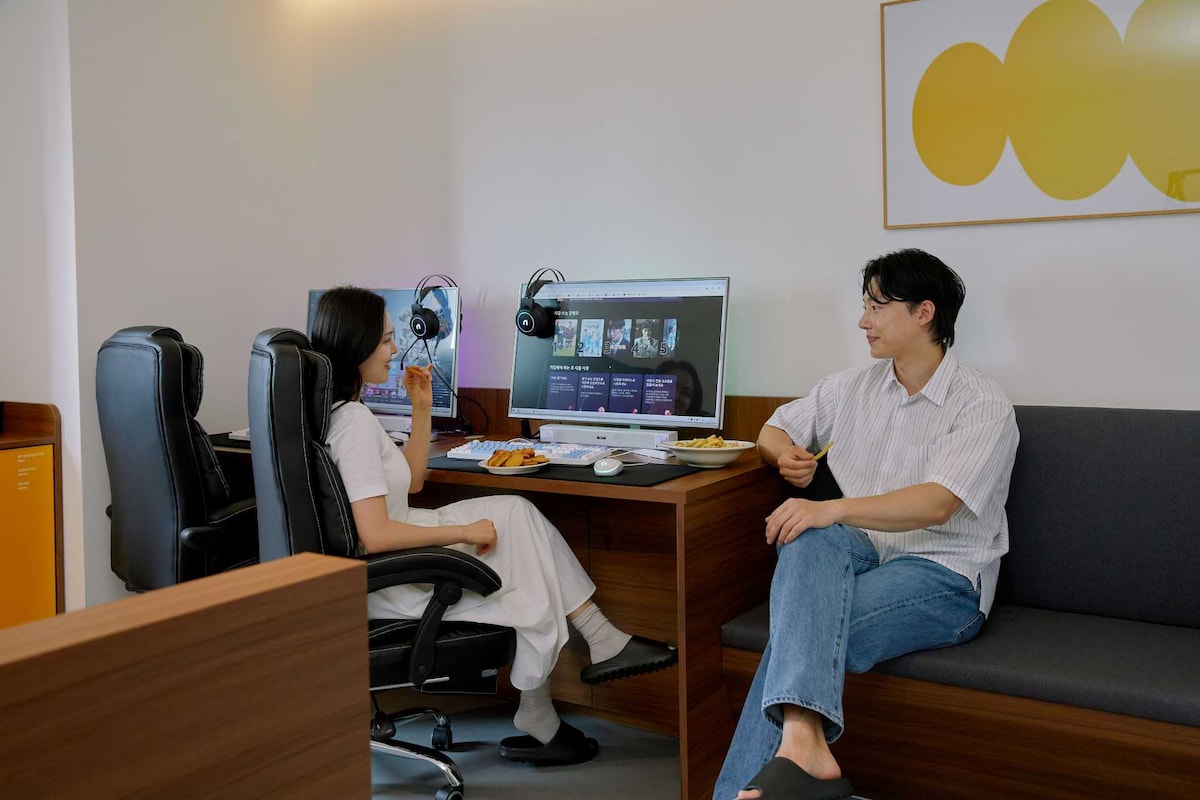 A modern workspace is showcased, featuring two comfortable chairs at a desk with a large computer screen. Headphones are placed on the desk. A cozy seating area with a plush couch is visible, providing a relaxed space for conversation.