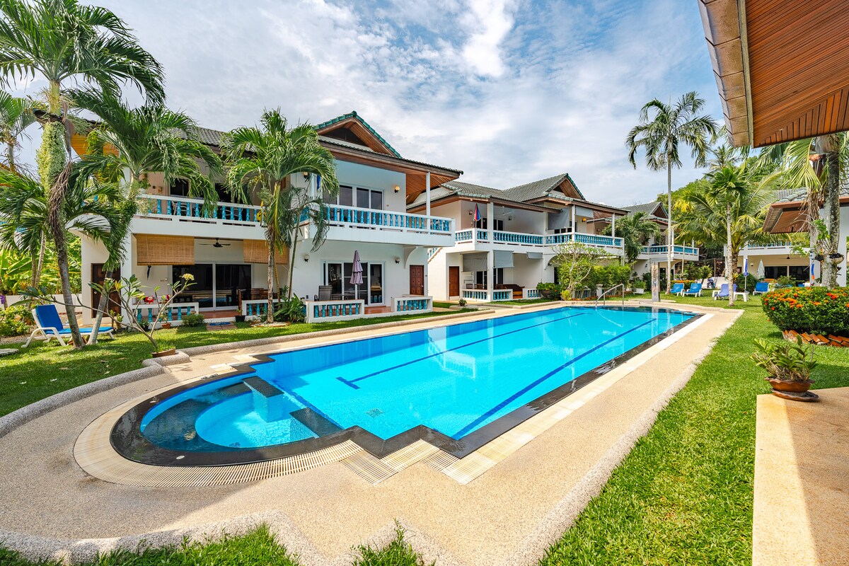 The image depicts a large, inviting swimming pool framed by lush green grass and tropical plants. Surrounding the pool, several buildings showcase balconies and patio areas with white railings. Sun loungers line the poolside, contributing to a serene and relaxed atmosphere.