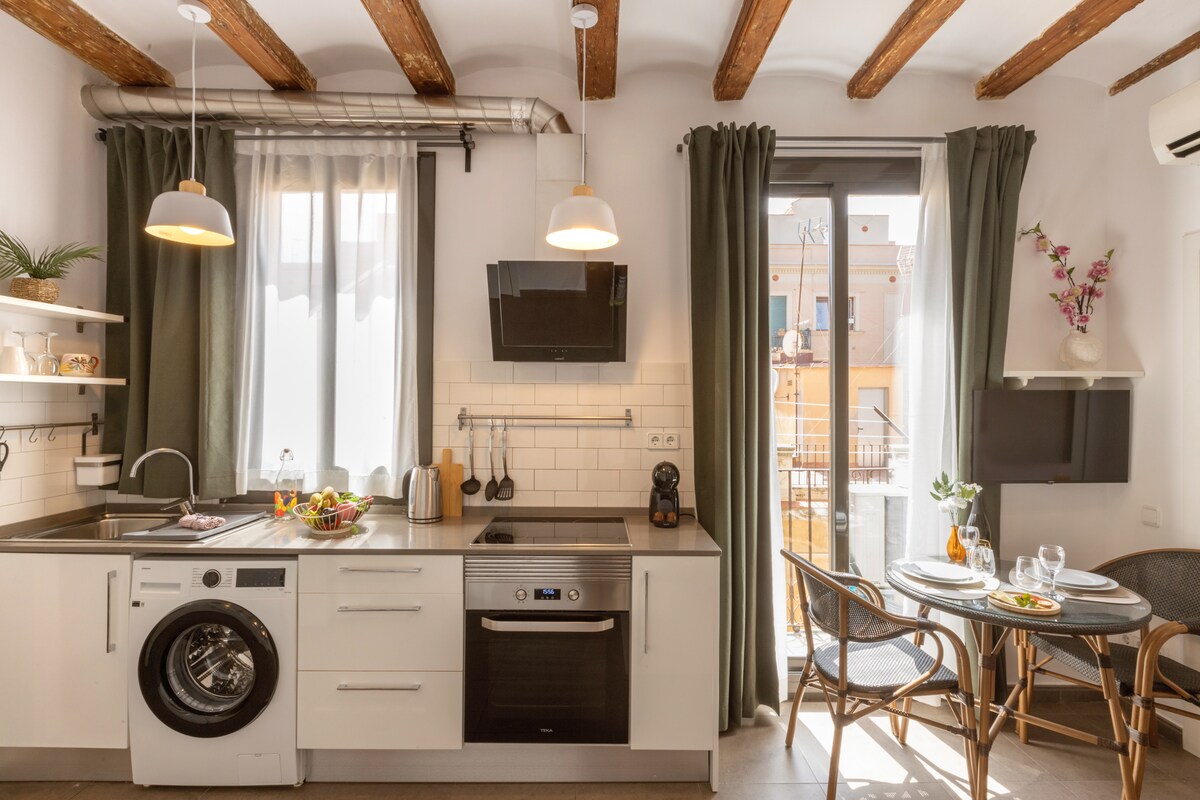 The modern kitchen features a washing machine and an oven, complemented by wooden beams overhead. Natural light flows through large windows framed by floor-length curtains, enhancing the inviting dining area with a round table set for two. A small balcony is visible beyond.