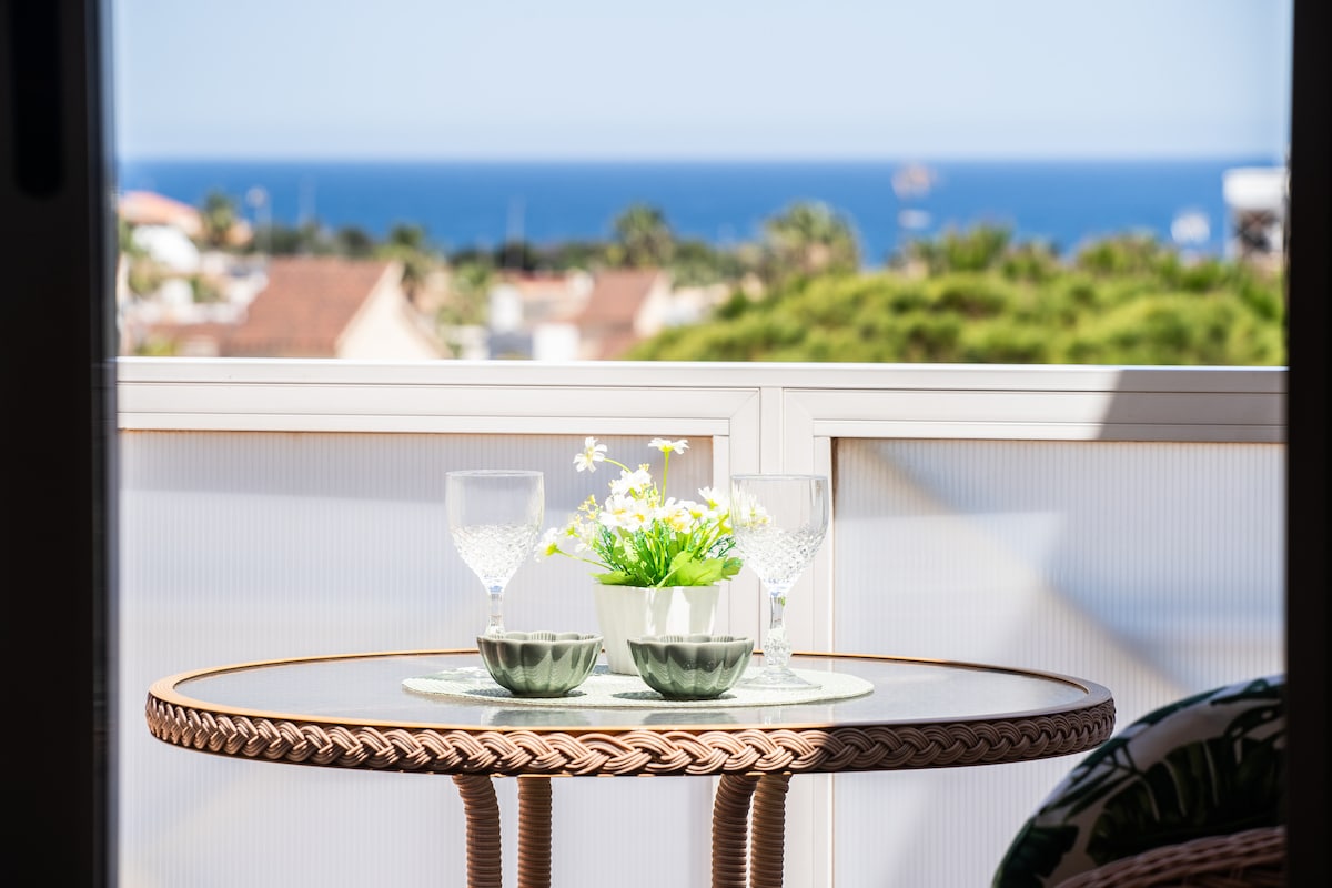 A small round table with a woven base is positioned on a balcony. It is set with two glasses and a decorative bowl holding fresh flowers. The background features a view of the blue Mediterranean Sea, enhancing the inviting outdoor space.
