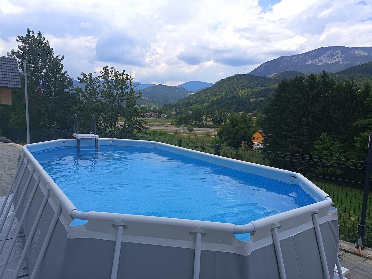 An above-ground pool is visible, surrounded by a paved area and overlooking a scenic landscape. The clear water reflects the sky, while mountains and greenery form a picturesque backdrop.