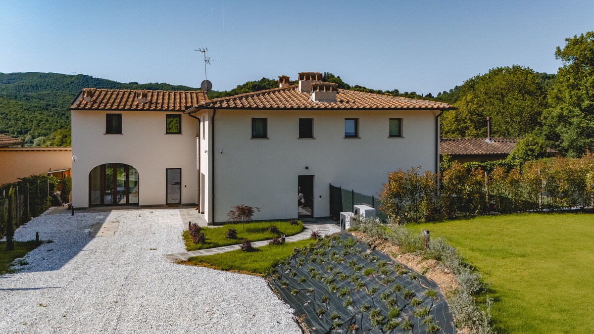 A two-story Tuscan farmhouse is depicted, featuring a terracotta tiled roof and a white exterior. A gravel pathway leads to the entrance, with neatly landscaped greenery surrounding the property. Rolling hills are visible in the background, enhancing the tranquil countryside environment.