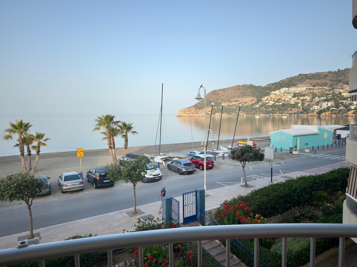 A view from the terrace showcases the calm sea and sandy beach, with palm trees lining the waterfront. Vehicles are parked along the street, and a colorful building can be seen nearby against the backdrop of a terraced hillside.