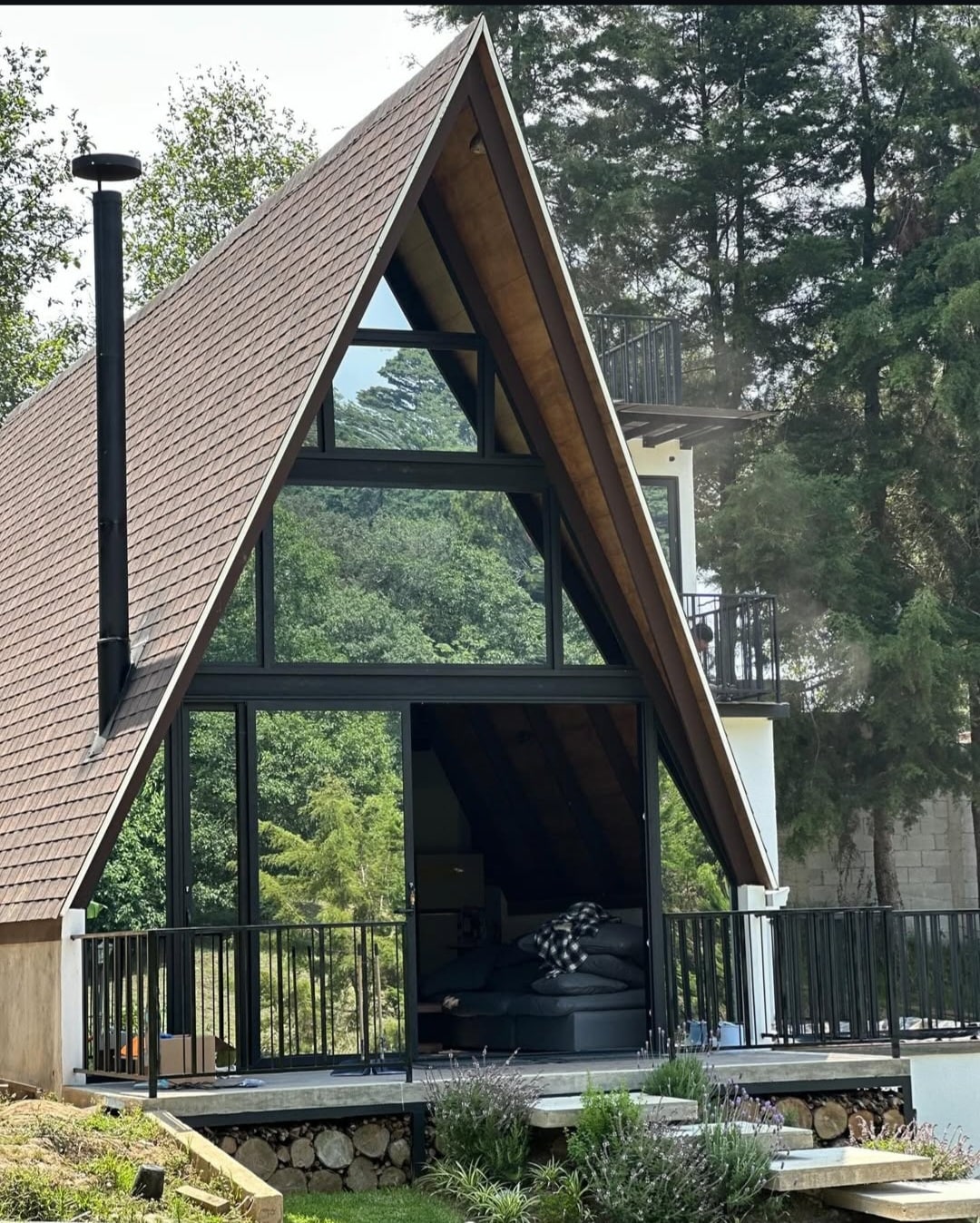 A modern A-frame cabin is surrounded by lush greenery, featuring large glass windows that invite natural light. The open balcony offers a view of the serene landscape, while a cozy seating area is visible inside, showcasing comfortable furnishings.