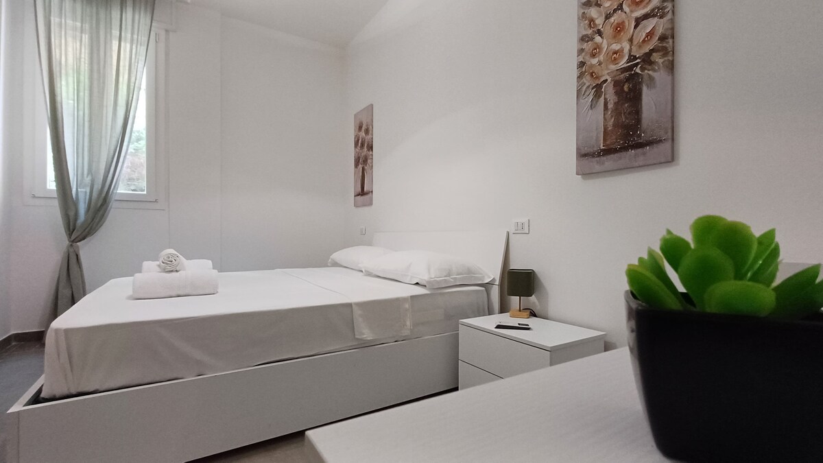 A bright bedroom features a spacious bed with white linens and a folded towel on top. A bedside table and a decorative plant are included, while artwork depicting flowers adorns the walls. Natural light enters from a window with sheer curtains.