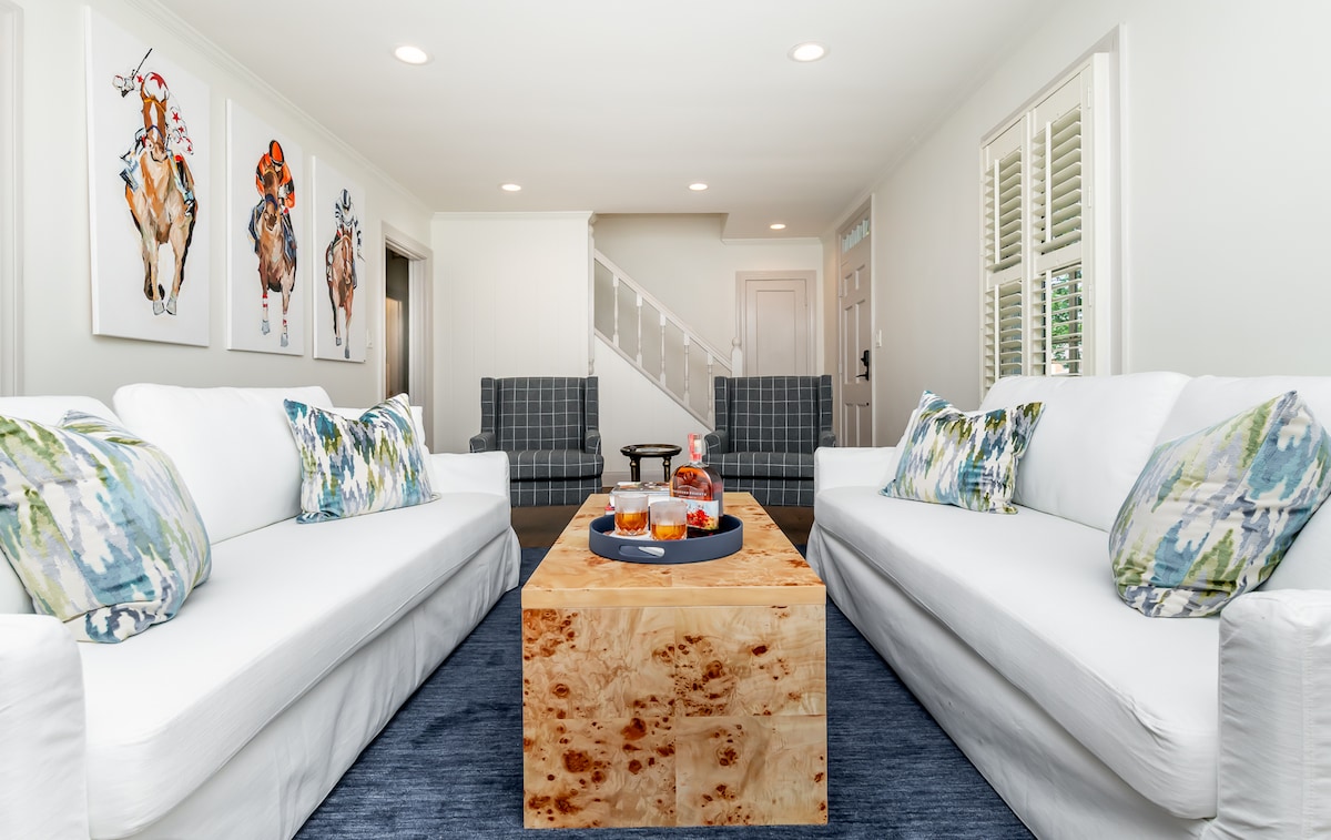 A bright living area features two white sofas, accented with colorful throw pillows. A rustic wooden coffee table sits centrally, displaying drinks. Two plaid armchairs are positioned across from the sofa, and vibrant artwork of cows adorns the wall, enhancing the inviting decor.