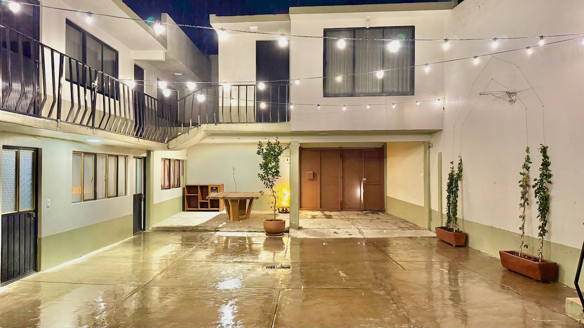 An open courtyard is featured, with polished concrete flooring reflecting ambient light. Strings of lights are stretched overhead, illuminating the space. A wooden table and benches are situated in one corner, while potted plants add a touch of greenery along the perimeter.