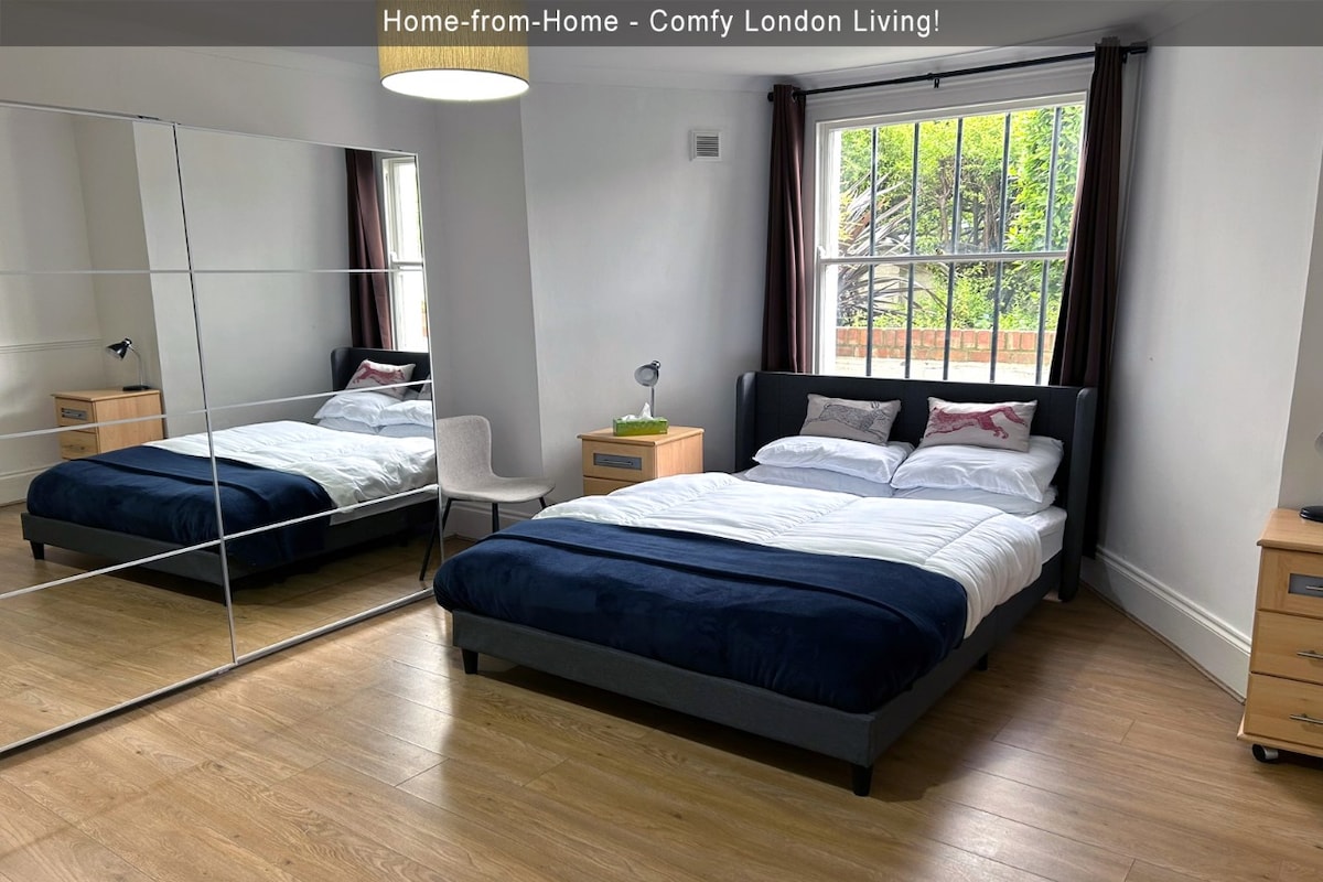 A spacious bedroom is highlighted by a large mirror, creating an airy feel. A comfortable bed is adorned with crisp linens and a navy throw. Natural light enters through two windows, framed by brown curtains, while a bedside table and lamp provide functional lighting.