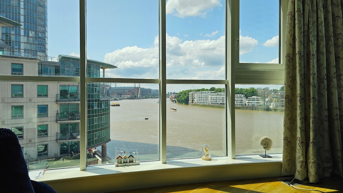 A panoramic view of the river Thames is framed by expansive floor-to-ceiling windows, allowing natural light to fill the space. Across the water, various boats are visible, with contemporary buildings lining the banks. The scene is complemented by decorative items on the windowsill.