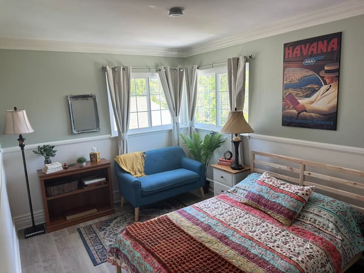 This upstairs bedroom features a full-size bed with a patterned quilt, two large windows, warm lighting, a reading nook with a loveseat, and storage space—great for relaxing after a day of exploring.