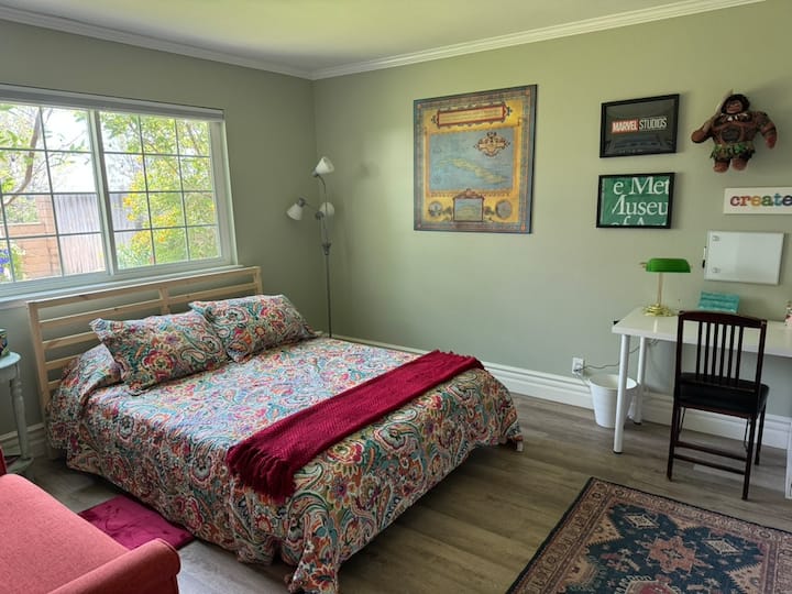 The downstairs bedroom features a queen bed with cozy, colorful bedding, plenty of natural light, a full-length window, and a writing desk—perfect for relaxing or getting a little work done during your stay.