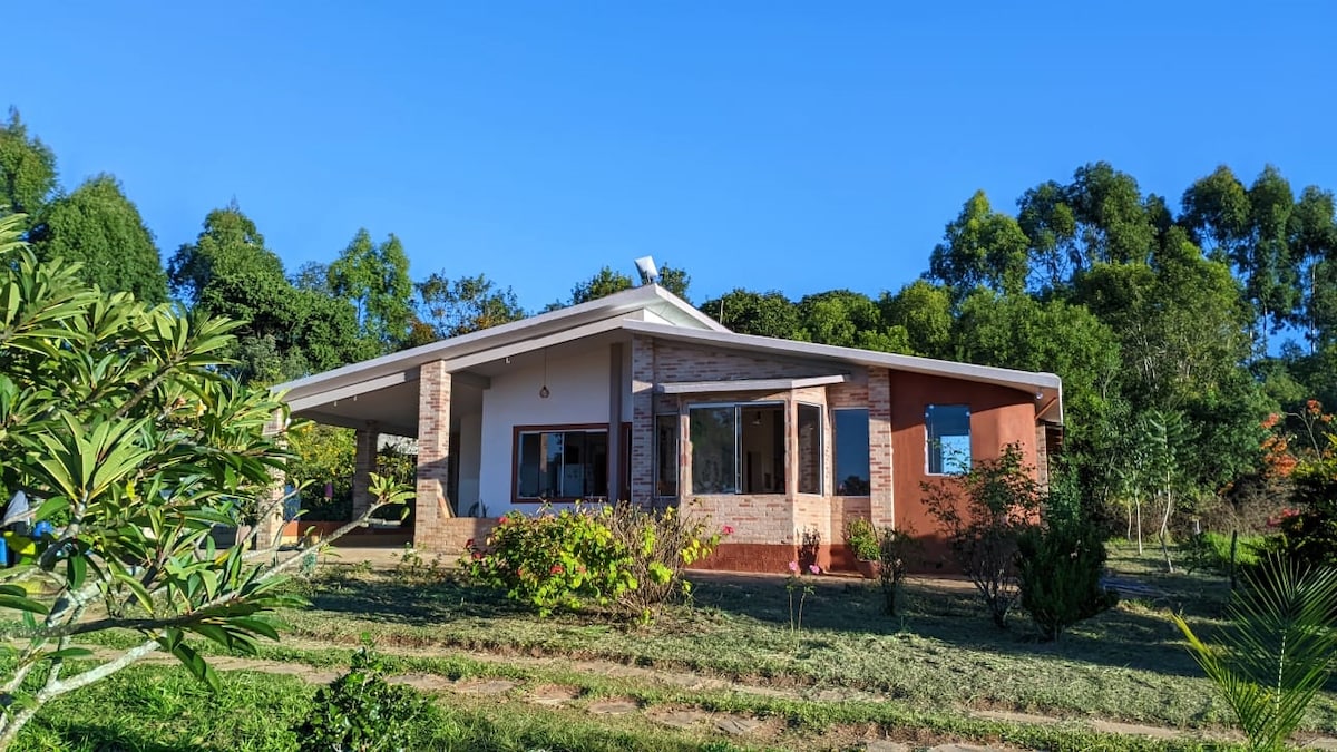 A spacious house is set amongst greenery, featuring a welcoming porch and large windows that allow natural light to fill the interior. The landscape includes well-maintained gardens and trees, contributing to a serene rural atmosphere.