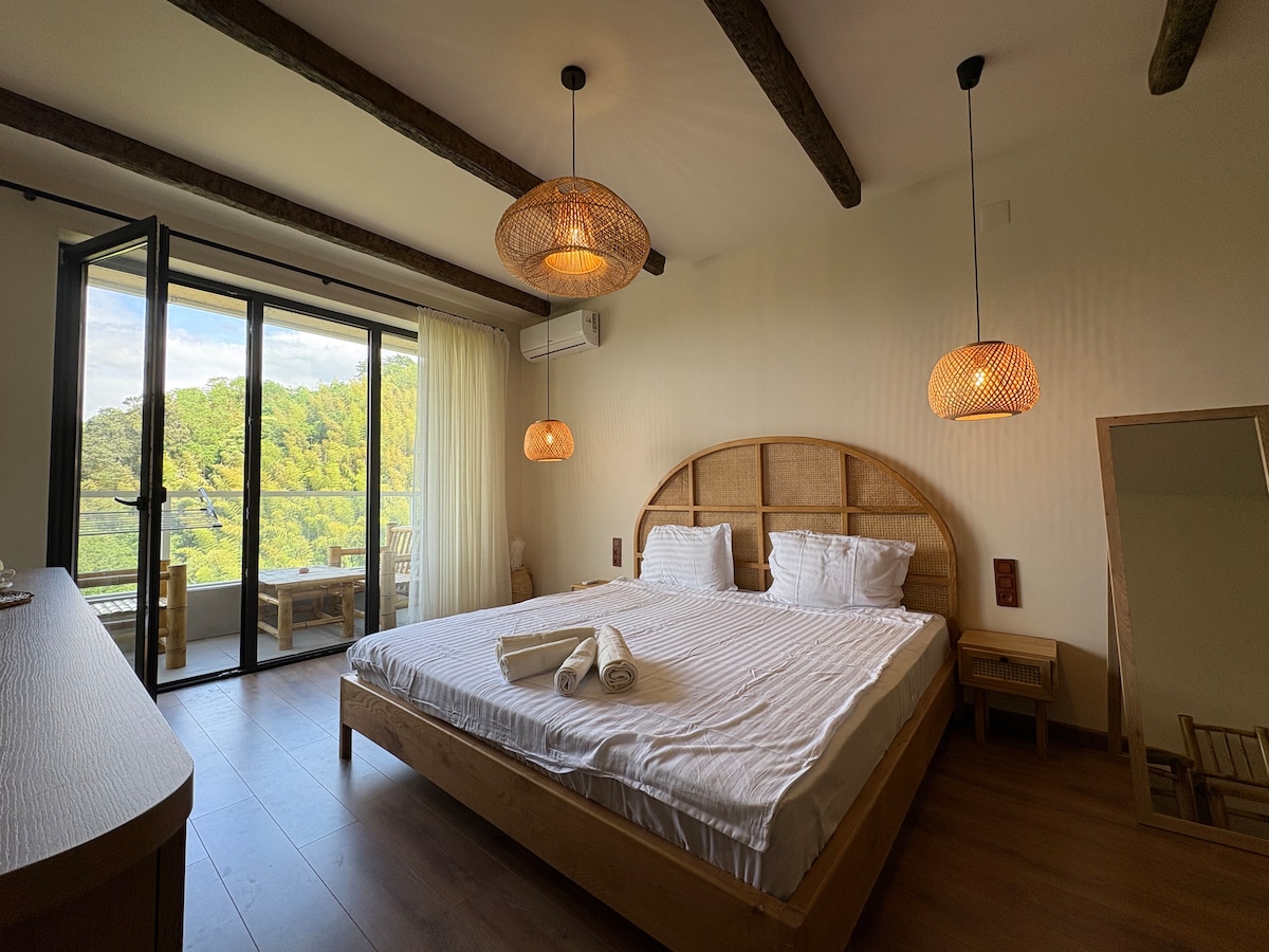 A well-lit bedroom is furnished with a spacious bed featuring crisp linens. Two woven pendant lights hang above. A patio door opens to a balcony with scenic views of greenery. A mirror and a wooden dresser complement the room's natural aesthetic.
