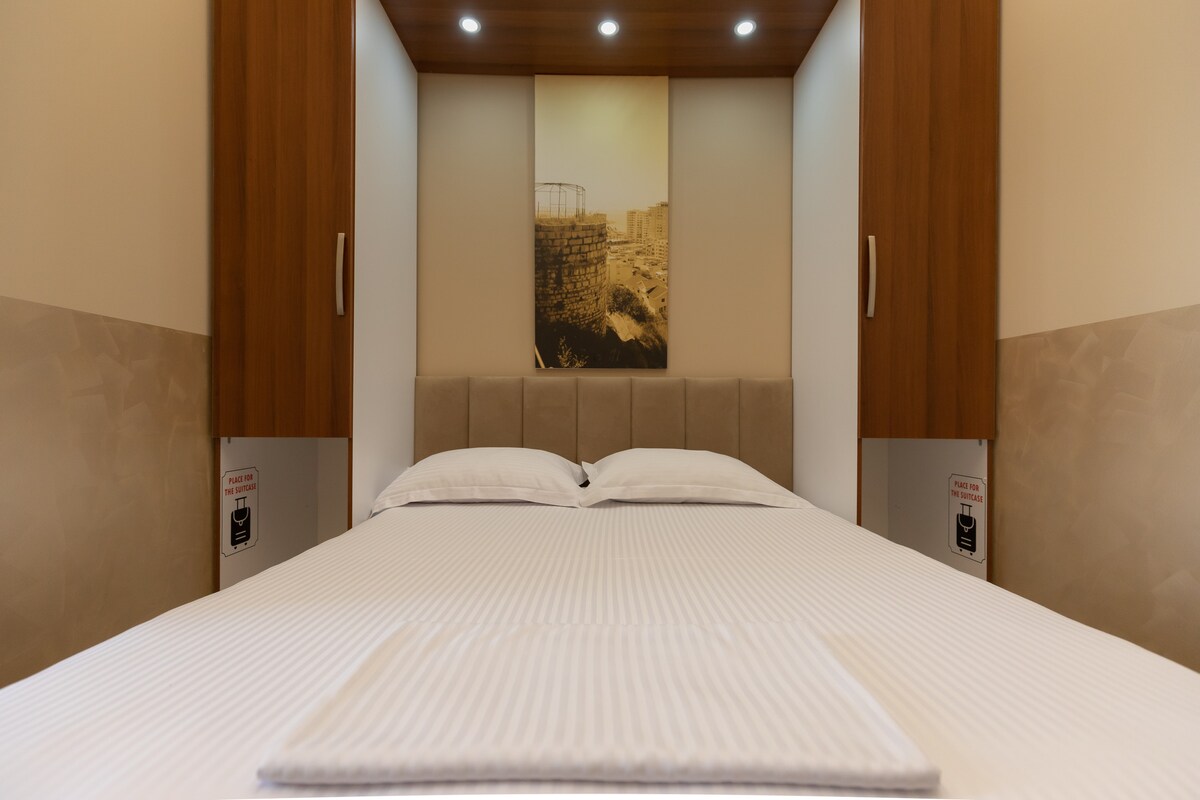A spacious bed is positioned centrally against a modern headboard within a minimalist sleeping area. Soft lighting is provided by recessed ceiling fixtures, highlighting a framed artwork that decorates the wall above. Nearby cabinetry features a smooth finish, contributing to a clean and organized aesthetic.