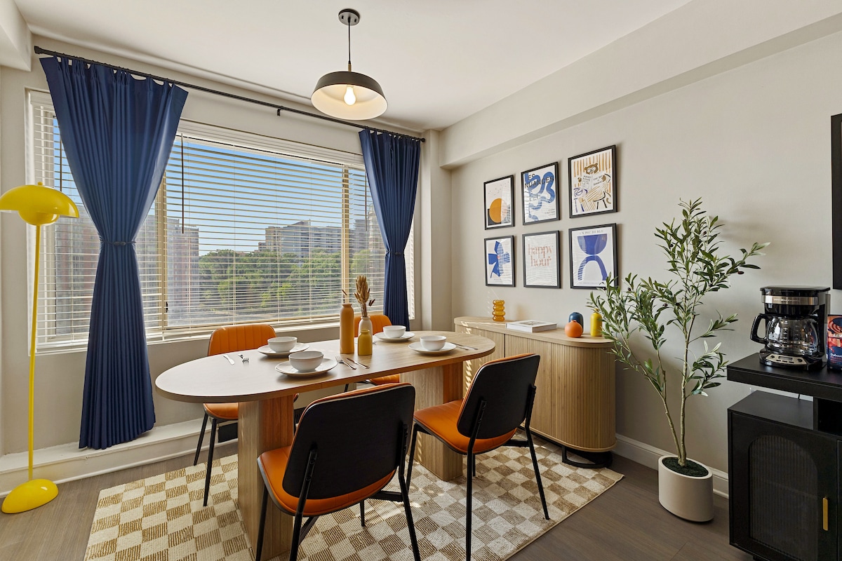 A dining nook features a round wooden table surrounded by four black chairs with orange cushions. Natural light filters through a large window adorned with blue curtains. Artwork hangs on the wall alongside a sideboard, while a coffee maker and decorative plants enhance the space.