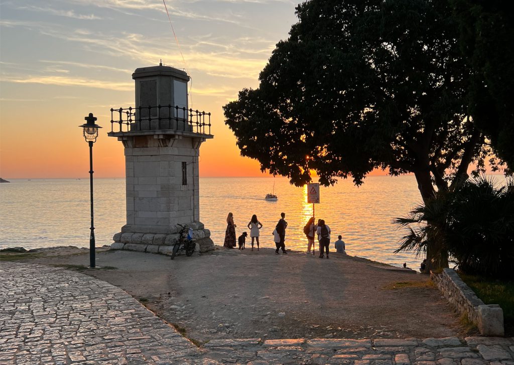 A tranquil waterfront scene at sunset features silhouettes of people enjoying the view. A lighthouse stands off to the side, with a lamp post adjacent to it. The calm sea reflects the warm hues of the setting sun, creating a serene atmosphere.