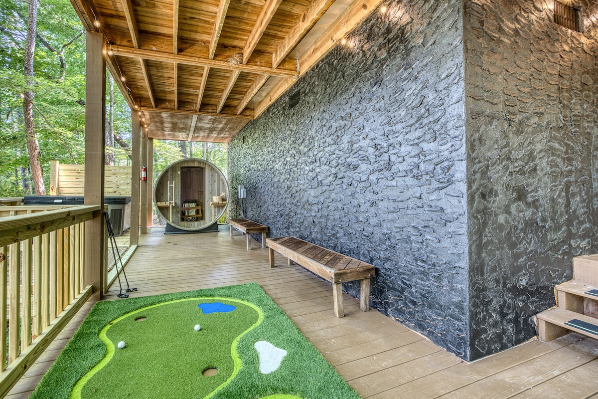 The outdoor area features a vibrant mini golf setup with an artificial green, complemented by a circular barrel sauna and wooden benches. Natural wood textures and stone walls create a serene backdrop, while string lights enhance the inviting atmosphere.