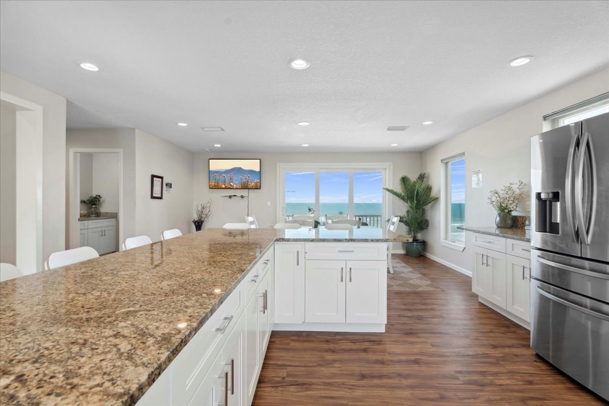 A modern kitchen is presented with a spacious granite countertop and white cabinetry. Stainless steel appliances are installed, while a large window provides ocean views. The open layout allows for easy flow into the dining area, with minimal decor enhancing a clean and functional space.