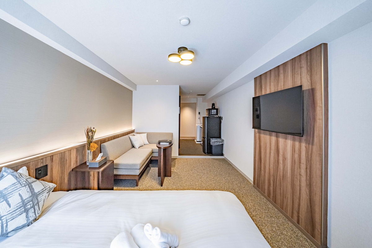 A bright and spacious room features a comfortable bed dressed in crisp white linens. A cozy seating area with a sofa is visible, accompanied by a small table. The room includes a flat-screen TV mounted on a wooden accent wall, providing a modern touch.