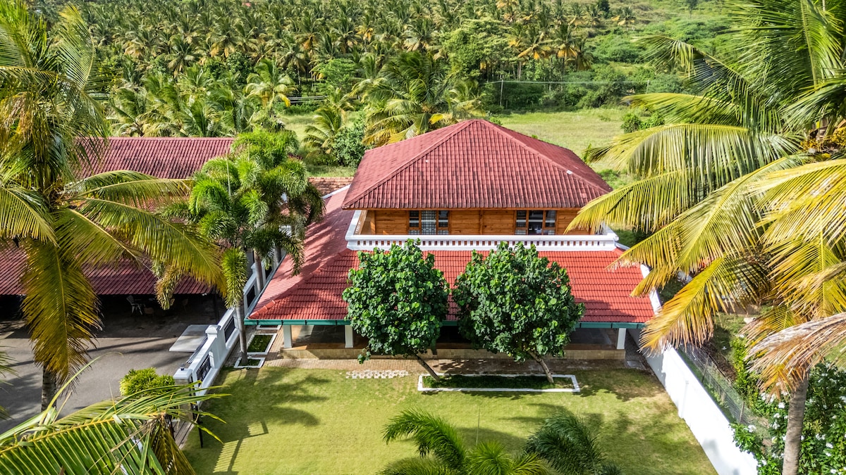 A spacious property is surrounded by lush greenery and tall palm trees. The structure features a red-tiled roof and is complemented by landscaped gardens. A clear view of the tranquil countryside can be seen in the background, providing a serene atmosphere.