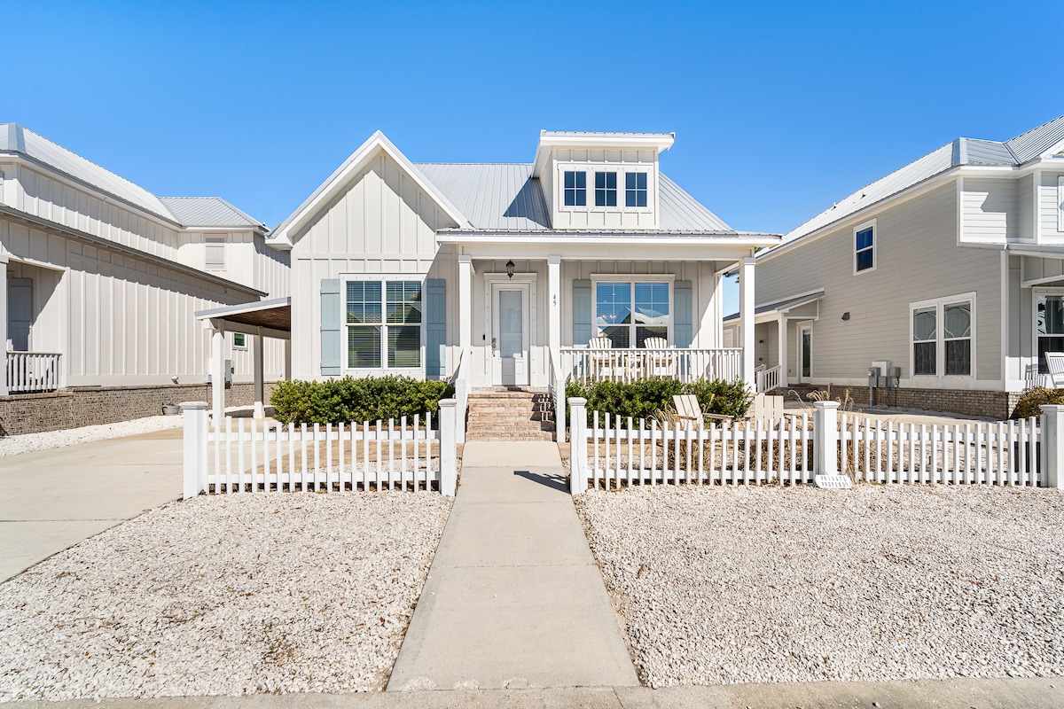 The exterior view of a coastal-style home is presented, featuring a light-colored facade with a peaked roof. A white picket fence encloses the front yard, which is landscaped with gravel and small shrubs. Clear blue skies provide a bright backdrop.