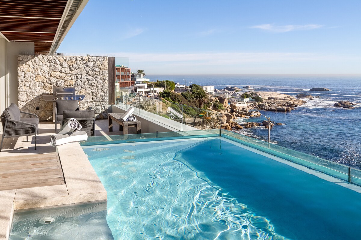 A private plunge pool with a transparent edge overlooks the ocean, offering stunning views of the coastline. Comfortable outdoor seating, including chairs and a gas BBQ, is positioned nearby on the spacious private terrace, enhancing the connection between indoor and outdoor living spaces.