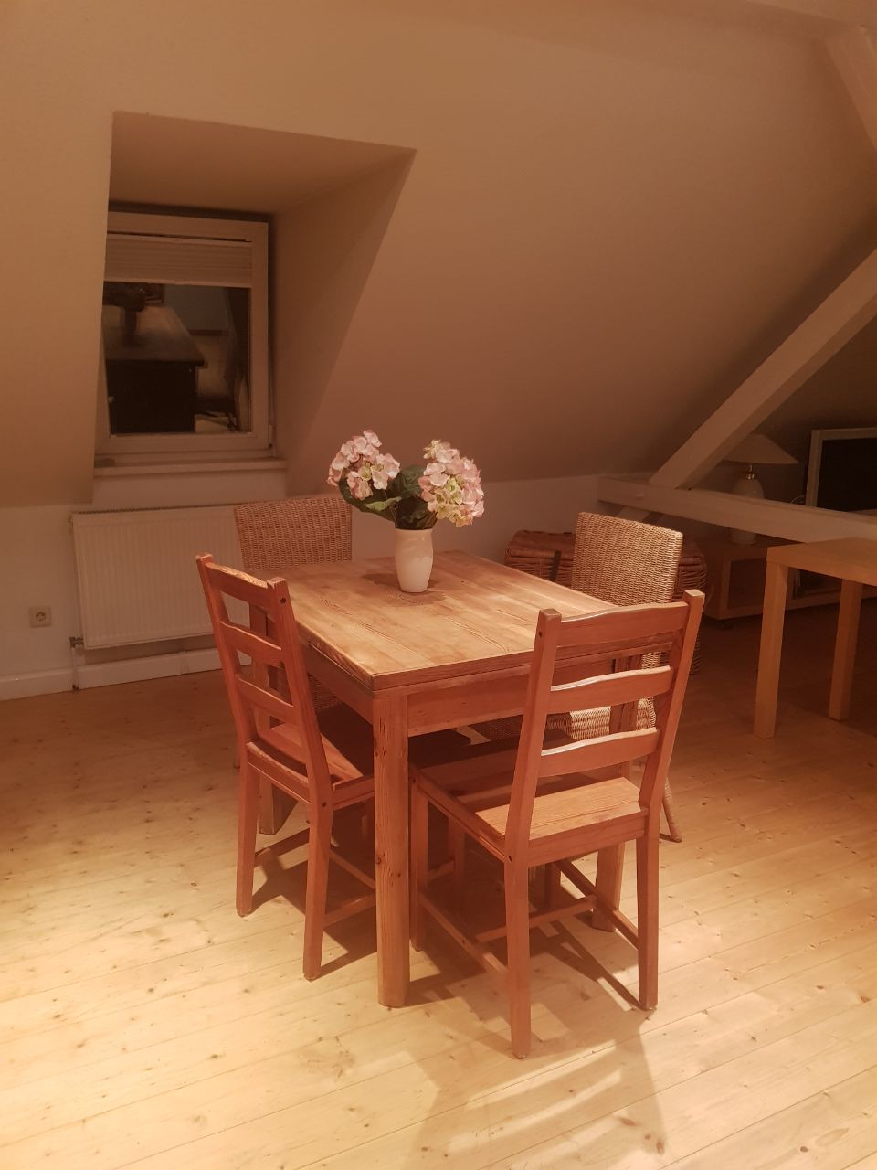 A wooden dining table is surrounded by four chairs, situated in a well-lit area beneath a sloped ceiling. A small vase with flowers is placed at the center of the table, adding a touch of decor. A window is visible in the background.