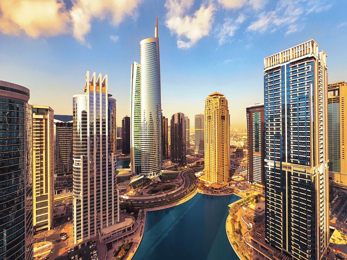 A skyline view features a mix of modern high-rise buildings surrounding a calm body of water. The architectural diversity includes glass facades reflecting sunlight, creating a dynamic cityscape under a blue sky dotted with clouds.
