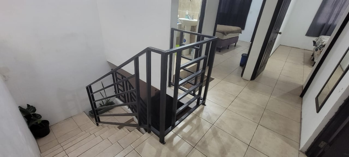 A staircase with a black metal railing is visible, leading to an upper level. The light-colored tiles reflect natural light coming from adjacent doorways. A hallway is seen, connecting to two separate rooms, one featuring a bed and a small seating area.