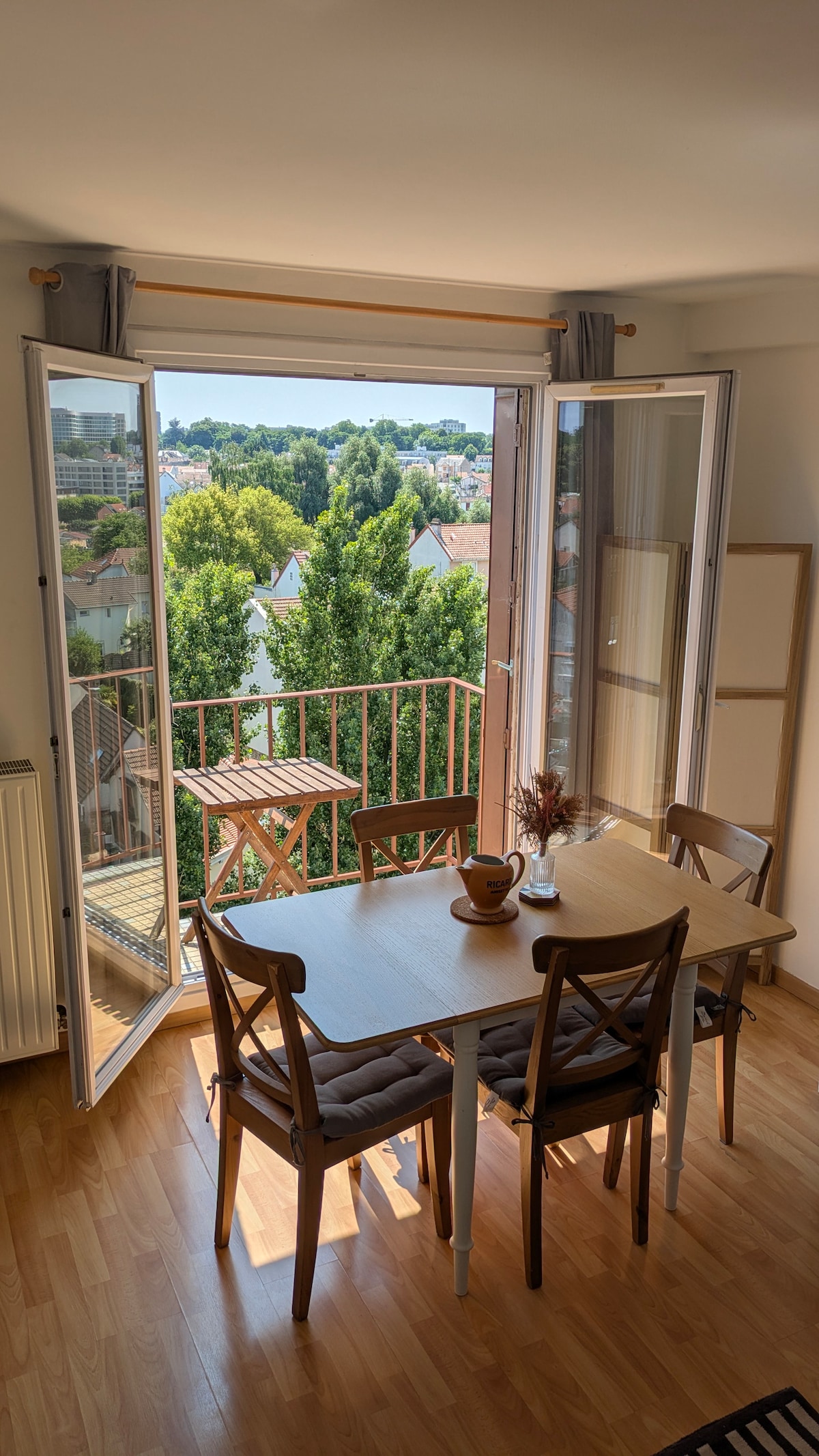 A dining area is equipped with a wooden table and four chairs, positioned near large open doors leading to a sunny balcony. Lush green trees and residential buildings are visible through the railing, creating a serene outdoor view.