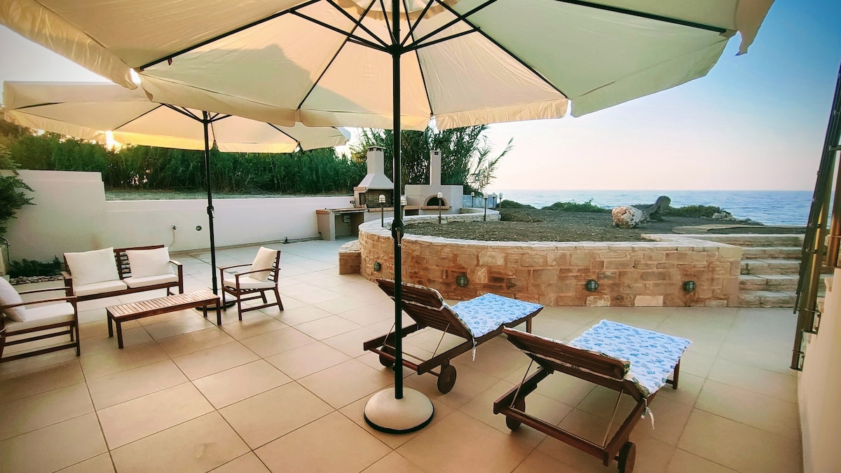 A spacious outdoor area features two sun loungers with patterned textiles, surrounded by shade provided by large umbrellas. A stone seating area is visible in the background, along with a grill. The coastline and natural landscape are seen beyond the terrace.