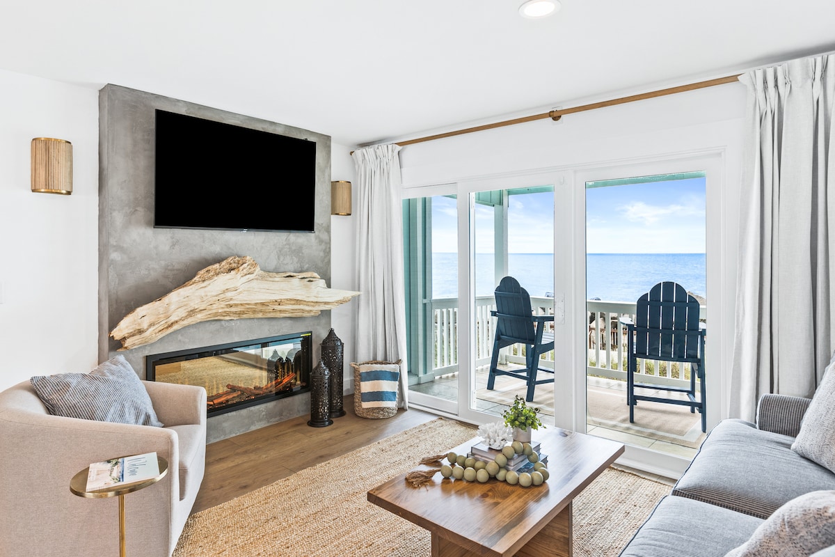 A cozy living area is adorned with a modern fireplace and a large television. Sliding glass doors lead to a private deck with ocean views, featuring two blue chairs. The space is highlighted by natural textures, including a neutral area rug and wooden accents.