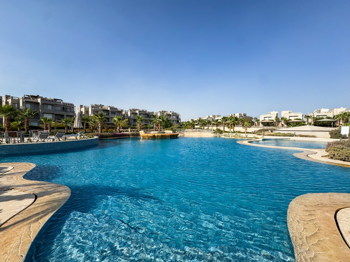 A large, crystal-clear swimming pool is surrounded by landscaped gardens and palm trees, offering multiple areas for relaxation. Sunny skies cast light on the water, reflecting shades of blue. Lounge chairs line the poolside, providing spaces for sunbathing and leisure.