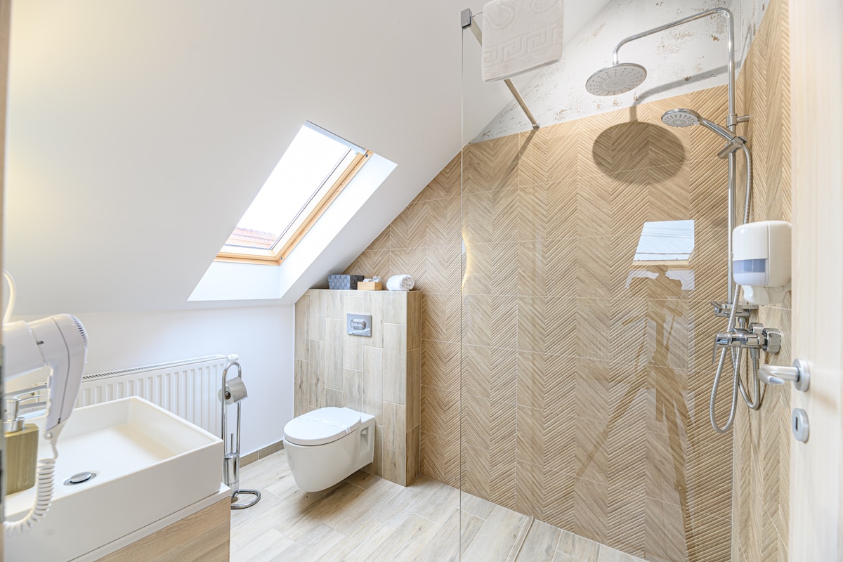 A modern bathroom features textured beige tiles with a herringbone pattern. A skylight allows natural light to illuminate the space. A wall-mounted toilet and a sleek wash basin with a faucet are provided, alongside a spacious shower area with dual shower heads.