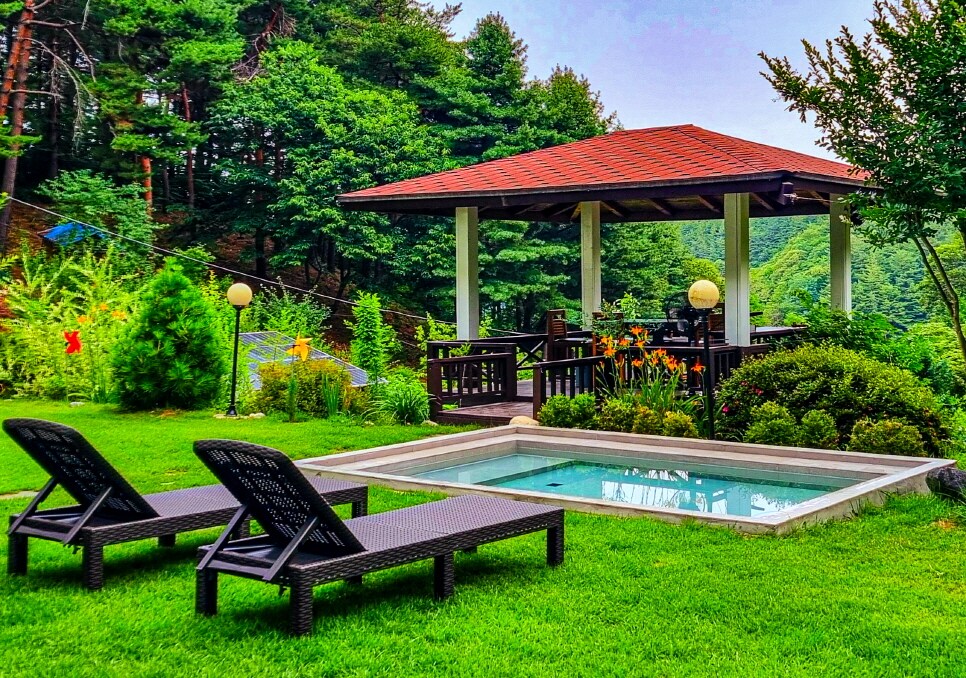 An outdoor area features a small pool surrounded by lush green grass and vibrant plants. Two lounge chairs are positioned nearby, offering a comfortable spot to relax. A traditional gazebo provides shade and seating, nestled among trees that enhance the natural setting.