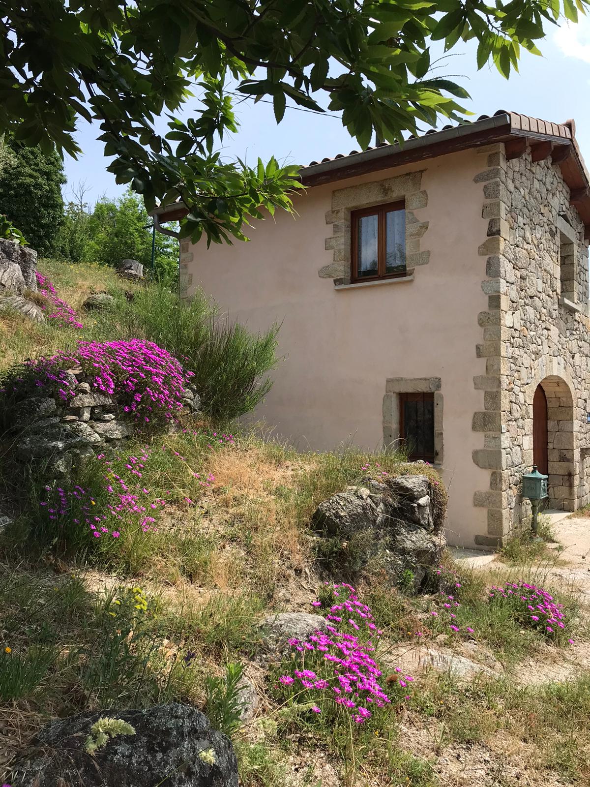 A charming stone cottage is surrounded by vibrant pink flowering plants and rocky terrain. The exterior features a warm, pastel wall and a wooden-framed window. The natural landscape provides a serene setting, with greenery in the background under a bright sky.