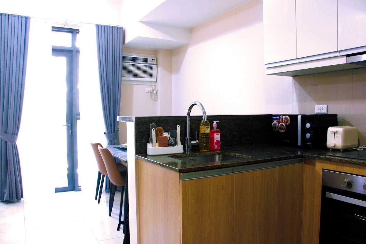 A modern kitchenette features sleek cabinetry and stainless-steel appliances, including a microwave and toaster. A small dining area is visible with four chairs surrounding a table. Natural light enters through the sliding glass door, which opens to a balcony, while light-colored curtains frame the window.