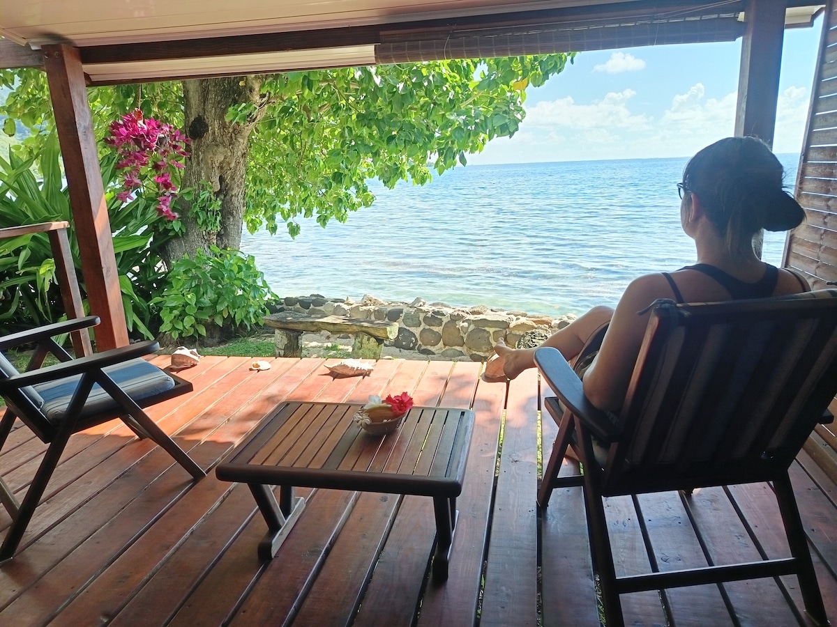 An outdoor seating area is visible, featuring a small wooden table and two chairs positioned on a wooden deck. A person sits relaxed, overlooking the calm ocean waters. Lush greenery and colorful flowers frame the scene, enhancing the natural beauty of the coastal view.