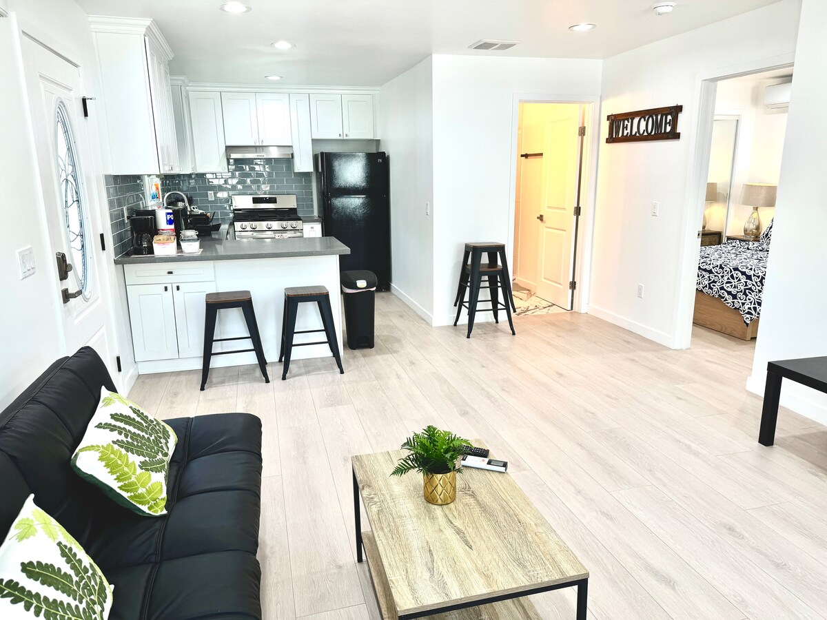 An open living area features a black sofa adorned with green cushions, and a light wooden coffee table with a small plant. A modern kitchen showcases white cabinetry and stainless steel appliances, with two stools positioned at the counter. Light-colored flooring enhances the spacious feel.