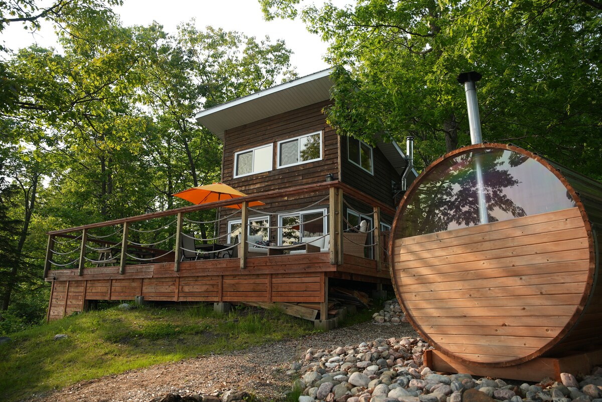 A cozy cabin is featured among lush trees, showcasing a spacious deck with several seating areas. A vibrant orange umbrella provides shade over outdoor seating. A wood-fired sauna, constructed with circular wooden planks, is positioned nearby, complemented by smooth stones lining the path.