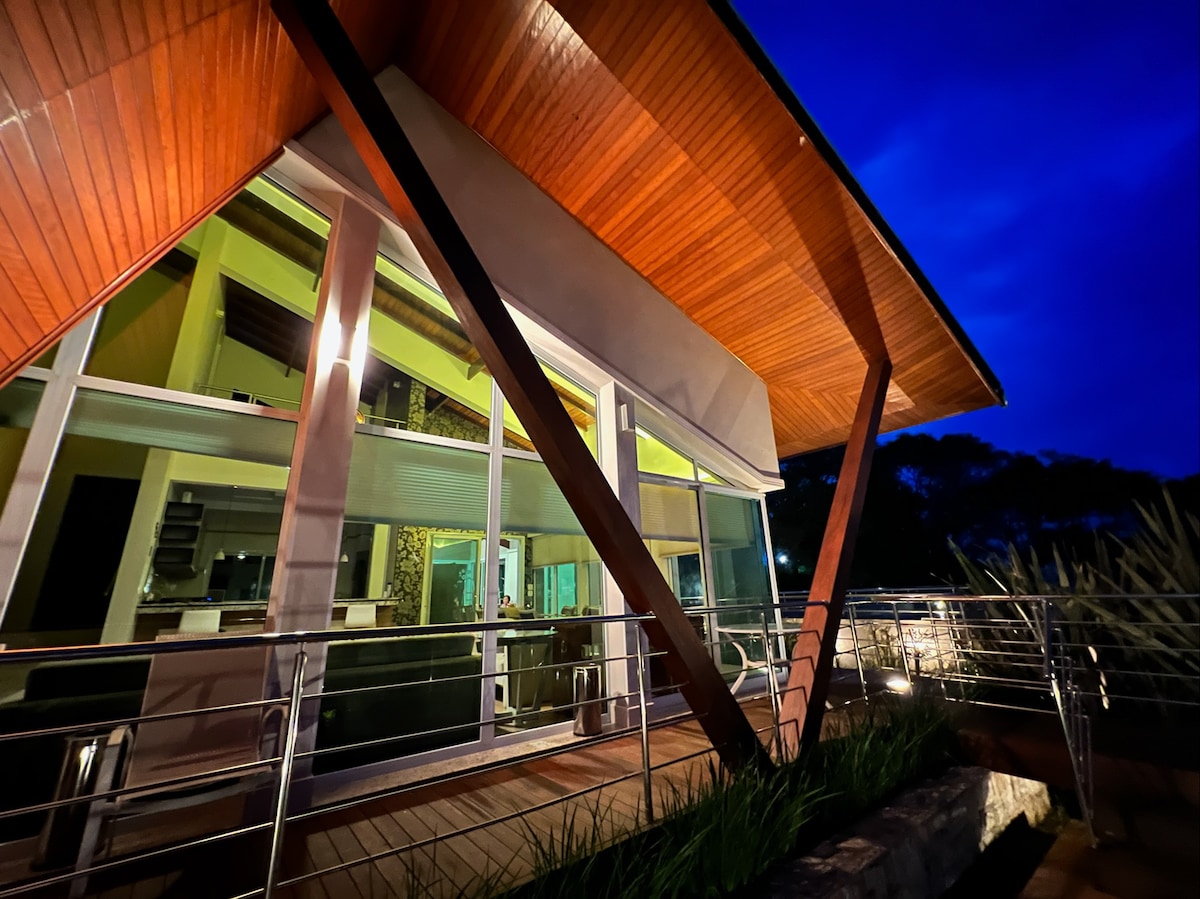 The modern structure features large glass windows and an angled wooden roof, accentuating its architectural design. Soft lighting from inside highlights the interior spaces, while a wooden deck and metal railing create a welcoming entrance. The surrounding greenery is subtly visible against a twilight sky.