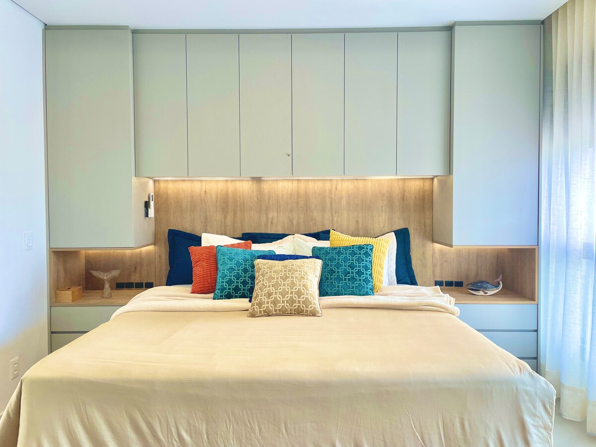 A spacious sleeping area features a large bed with a light-colored duvet, adorned with a variety of colorful pillows. The headboard is complemented by illuminated panels, and built-in cabinetry offers additional storage. Soft natural light filters through the sheer curtains, enhancing the serene atmosphere.