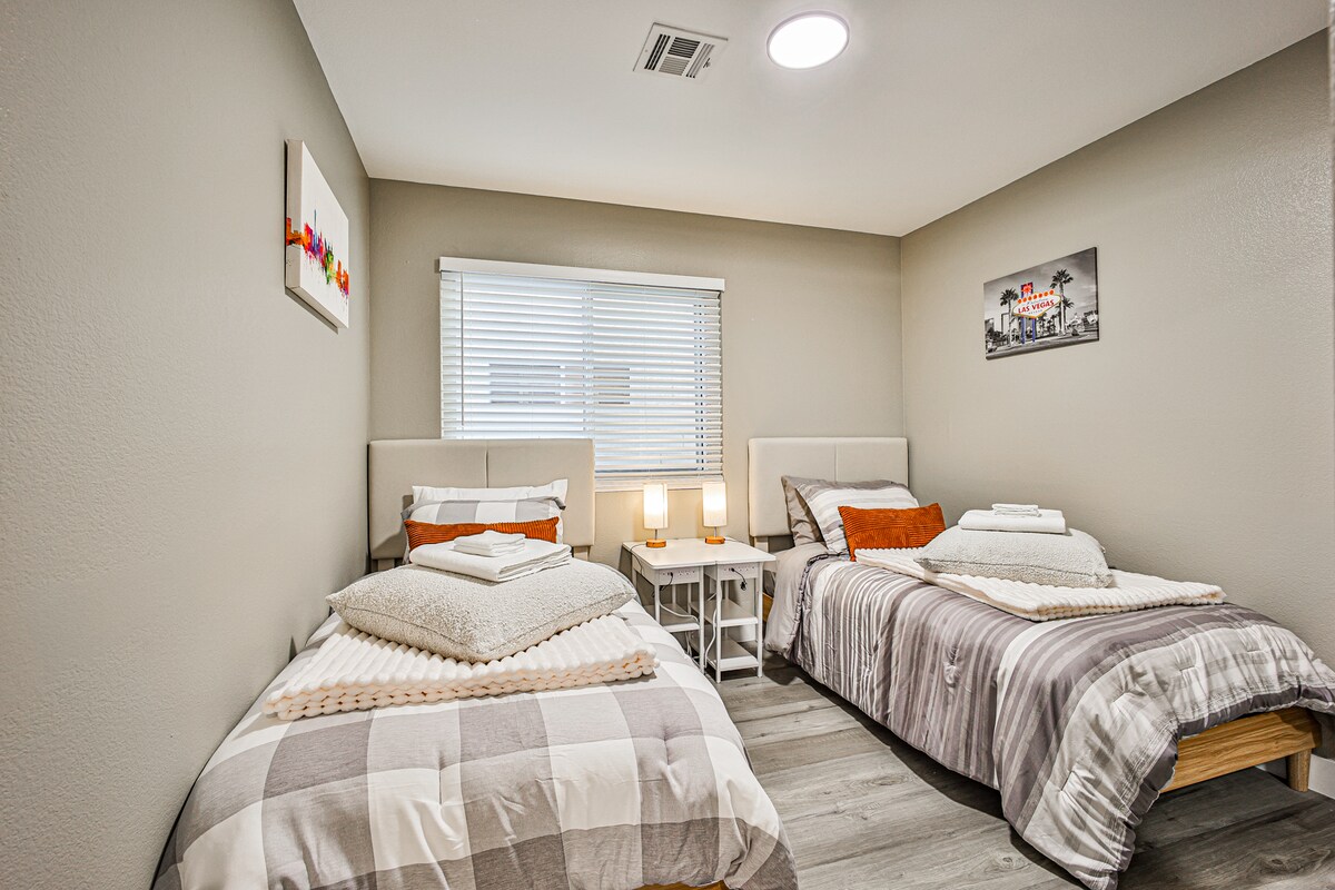 Deze gezellige slaapkamer in The Vegas Hideout heeft twee eenpersoonsbedden met zacht beddengoed, oranje sierkussens, een gedeeld nachtkastje met lampen en kunst aan de muur. Lichte houten vloeren en een raam met jaloezieën maken het licht, schoon en ideaal voor langere verblijven.