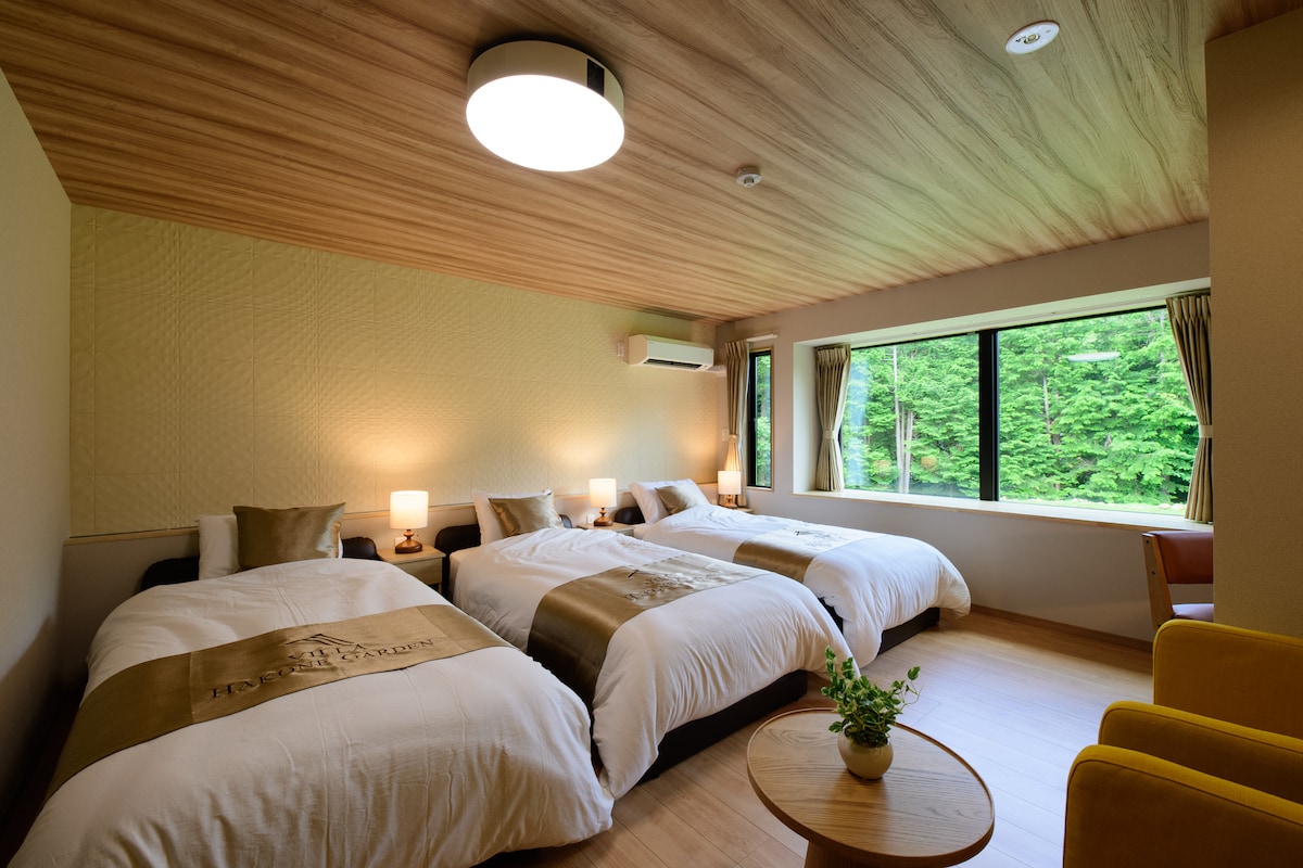 A well-appointed room features three spacious beds with decorative throws. Soft lighting is provided by bedside lamps, and large windows offer views of the surrounding greenery. A small table with a potted plant adds a touch of nature indoors, enhancing the tranquil ambiance.