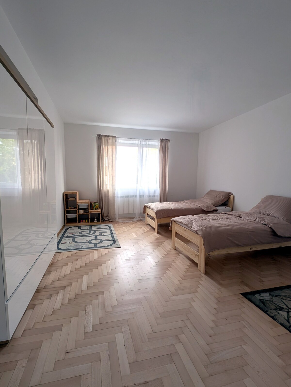 A lightly furnished room features two wooden single beds with neutral bedding positioned near a large window, allowing natural light to brighten the space. A mirrored wardrobe and shelving unit offer storage options, and two area rugs enhance the wooden flooring.