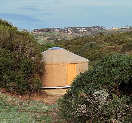A yurt with a rounded structure is nestled among dense greenery. The exterior is a soft tan color, with a wooden door and a circular skylight on top. The surrounding landscape features rolling hills in the background, contributing to a serene and secluded atmosphere.