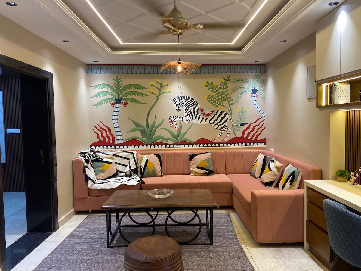 A spacious living area features an L-shaped coral sofa adorned with decorative cushions. A wooden coffee table sits in the center on a textured rug. The wall showcases a vibrant mural of tropical flora and fauna, enhancing the room's relaxed ambiance.